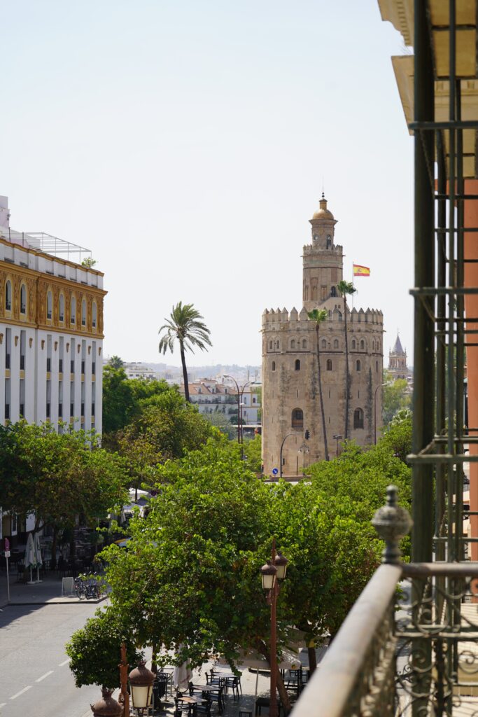 Vista de la Torre del Oro desde una ventana con rejería verde de Tayko Sevilla, entre palmeras y árboles