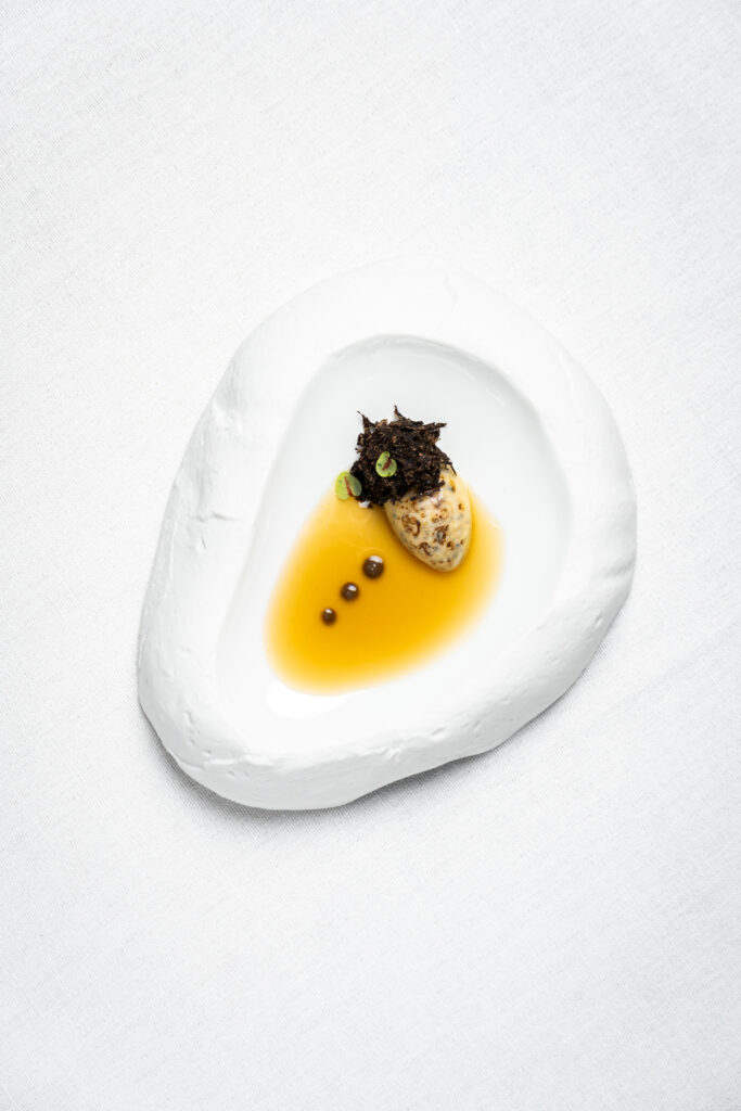 A haute cuisine dish seen from above: golden sauce on an organically shaped white plate, with an omelette, a dark garnish of green shoots and three small pearls, on a white tablecloth.